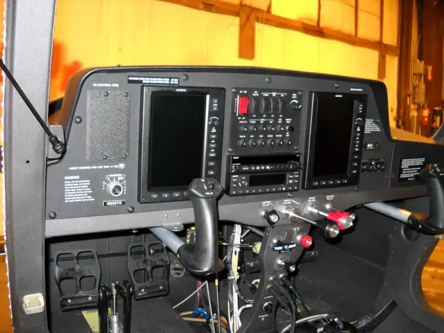 A light aircraft instrument panel with twin Garmin glass displays, photographed in a hangar with wiring exposed beneath it.